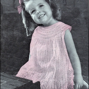 May include: A little girl wearing a pink crocheted dress with a pink bow in her hair. The dress has a scalloped hem and a crocheted bodice. The girl is smiling and looking up.