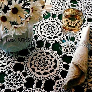 Puede incluir: Mantel de encaje blanco con un delicado patrón floral. Un vaso de vino y una servilleta blanca se colocan sobre el mantel. El texto "Tick Tock Knits" es visible en la esquina inferior izquierda de la imagen.