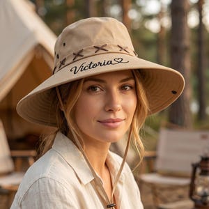 May include: Tan sun hat with a wide brim and a chin strap. The hat features brown leather-like accents and the name "Victoria" in black script. The hat is designed for outdoor activities and sun protection.