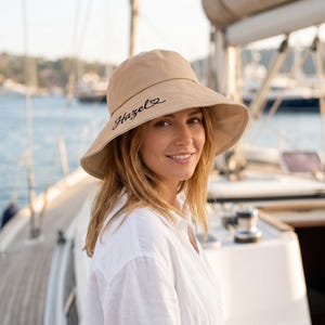 Könnte beinhalten: Ein beiger Sonnenhut mit breiter Krempe. Der Hut trägt den Namen "Hazel" in schwarzer Schrift. Die Person auf dem Bild trägt ein weißes Hemd mit Knöpfen und befindet sich auf einem Boot.