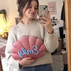 May include: A plush, shell-shaped cushion in a dusty rose colour featuring the name Emily embroidered in a light blue, textured script font across the centre. The cushion has a distinct scalloped design with stitched vertical lines radiating outward.
