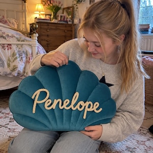 Op de afbeelding: Een blauwgroen fluwelen kussen in de vorm van een schelp met de naam Penelope in crèmekleurig touwschrift in het midden. Het kussen heeft een opvallend geschulpt randontwerp met verticale lijnen die vanuit de basis naar boven lopen.