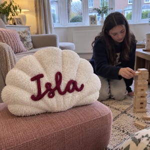 May include: A white, textured, shell-shaped decorative cushion featuring the name Isla embroidered in a dark red, chunky yarn script across the centre. The cushion has a soft, plush surface with distinct ridges radiating from the bottom edge.
