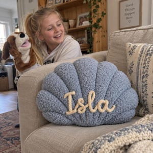 Könnte beinhalten: Ein staubblaues, muschelförmiges Dekokissen mit einer strukturierten, boucléartigen Oberfläche. Der Name Isla ist in einer cremefarbenen, kursiven Seiloptik-Schriftart in der Mitte des Kissens eingestickt, das auf einem beigen Sofa liegt.