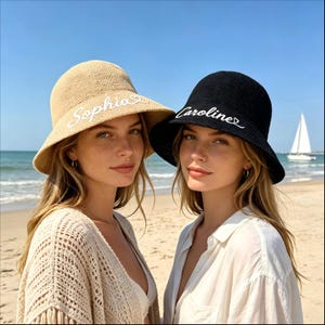 Könnte beinhalten: Zwei Frauen am Strand, jede mit einem Sonnenhut. Einer ist beige mit weiß gesticktem "Sophia", der andere ist schwarz mit weiß gesticktem "Caroline". Der Hintergrund umfasst das Meer und ein Segelboot.