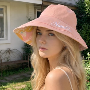 May include: A peach-colored bucket hat featuring the word Name embroidered in white cursive script on the front. The underside of the wide brim displays a floral pattern in shades of yellow, white, and pink, visible from the front angle.