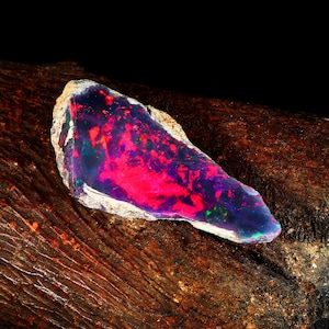 May include: A colourful, raw opal gemstone. The opal displays a deep purple base with fiery red and green flashes. The stone is set against a dark, textured background. The opal is a natural, uncut stone.