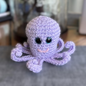 May include: A light purple crocheted octopus plush toy featuring large, round, glossy black eyes with blue iris accents and a small, embroidered pink mouth. The octopus has a rounded head and eight curled, textured tentacles extending outward.