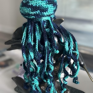 May include: A crocheted jellyfish toy featuring a variegated yarn pattern in shades of dark navy blue and bright turquoise. The jellyfish has a rounded, dome-shaped head and numerous long, spiraling, dangling tentacles of varying lengths.