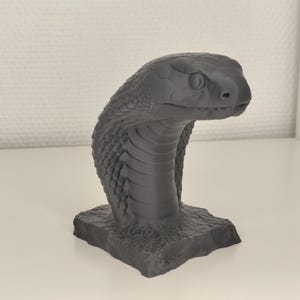 May include: A grey cobra sculpture with a detailed, textured surface. The snake's head is raised, and the sculpture is mounted on a rough, stone-like base. The cobra's scales and facial features are clearly defined.