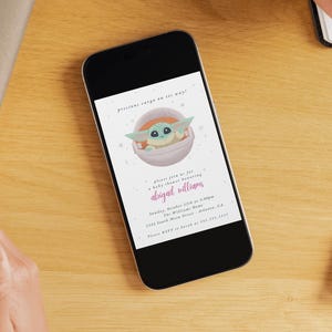 May include: A smartphone displays a baby shower invitation with a cartoon character in a pod. The invitation text reads "precious cargo on its way!" and includes the name "abigail williams" in pink script. The phone rests on a wooden surface.