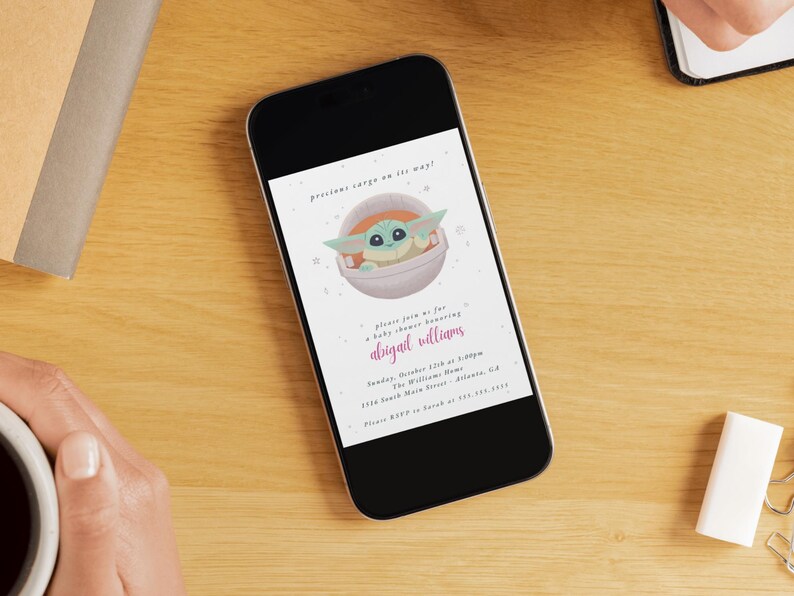 May include: A smartphone displays a baby shower invitation with a cartoon character in a pod. The invitation text reads "precious cargo on its way!" and includes the name "abigail williams" in pink script. The phone rests on a wooden surface.