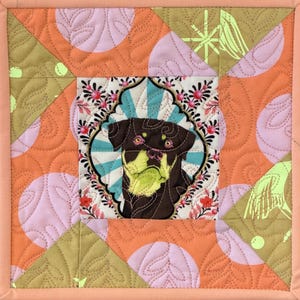 May include: A square, quilted wall hanging featuring a dog portrait. The dog is brown and yellow, set against a floral and geometric pattern in shades of orange, pink, and green. The piece is framed with a light peach border.