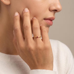 May include: A gold ring with a small, round, red gemstone. The ring is on a person's finger, with the hand and part of the face visible. The ring is simple and elegant.