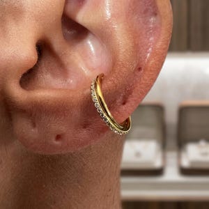 May include: A close-up of a gold hoop earring with small, clear gemstones set along the outer edge. The earring is worn in the ear of a person. The earring is a simple, elegant design.