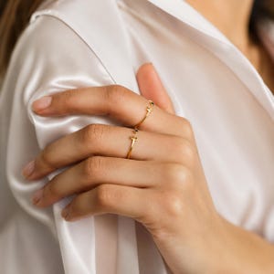 Puede incluir: Dos delicados anillos dorados con pequeñas piedras preciosas se llevan en una mano. Los anillos tienen un diseño minimalista, con una banda fina y un pequeño acento de piedra preciosa. La mano está colocada frente a una tela de satén blanca.