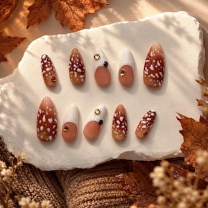 Op de afbeelding: Een set van tien opplaknagels in amandelvorm. De ontwerpen bevatten een bruin kleurverloop met witte spikkels, effen witte punten op een nude basis, en accenten van kleine gouden en zwarte ronde steentjes op geselecteerde nagels.