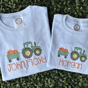 Puede incluir: Dos camisetas azul claro con un tractor verde bordado que tira de un remolque lleno de calabazas naranjas. La camiseta de la izquierda tiene bordado el nombre John Floyd en naranja, y la de la derecha tiene el nombre Morgan en naranja.