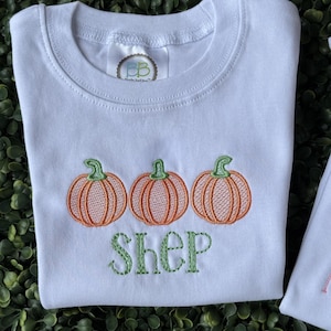 Puede incluir: Una camiseta blanca de manga corta con un diseño bordado de tres calabazas naranjas con tallos verdes. Debajo de las calabazas, el nombre SHEP está bordado en letras mayúsculas verdes. Una etiqueta de marca BB es visible en el cuello.