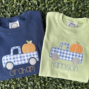 May include: A navy blue t-shirt and a light green t-shirt, each featuring an embroidered blue and white gingham truck carrying an orange pumpkin. The navy shirt has the name Graham embroidered in orange, and the green shirt has the name Harrison embroidered in blue.