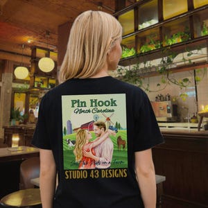 May include: Black t-shirt with a graphic on the back featuring a couple embracing in front of a farm scene. The graphic includes the text "Pin Hook North Carolina" and "Just Two Nuts on a Farm" with "STUDIO 43 DESIGNS" below.