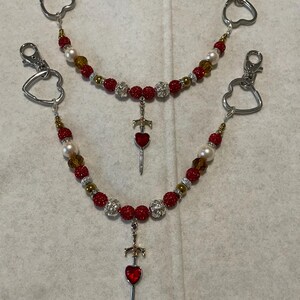 May include: Two beaded keychains with silver-tone heart-shaped clasps. The chains feature red, gold, and pearl-colored beads, with a sword pendant and a red heart charm. The keychains are designed to be attached to keys or other items.