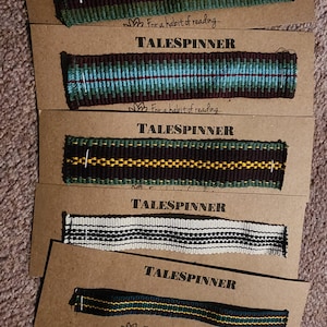 May include: Five woven bookmark bands in various color combinations, including green, brown, blue, and black, with yellow and white accents. Each bookmark is displayed on a brown card with the text "TALESPINNER" and "For a habit of reading..."