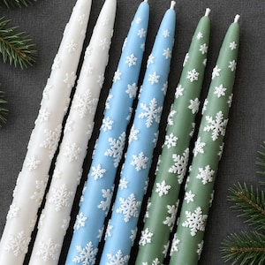 Peut inclure: Un ensemble de six bougies effilées ornées d'un motif de flocons de neige blancs en relief. Les bougies sont disposées par paires de couleurs blanc, bleu clair et vert sauge, avec une extrémité pointue et une base plate pour bougeoirs.