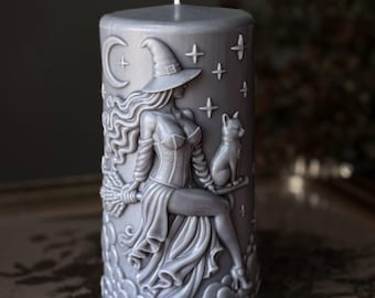 Puede incluir: Una vela cilíndrica de color gris con un diseño en relieve de una figura con sombrero puntiagudo sentada sobre una escoba, acompañada por un gato. La superficie incluye detalles grabados de una luna creciente, estrellas y nubes.