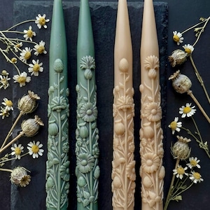 Peut inclure: Quatre bougies coniques décoratives dans les tons de vert et beige, finement sculptées de motifs floraux. Les bougies sont disposées sur une surface d'ardoise foncée, avec des fleurs séchées et des capsules de graines éparpillées. Les bougies mesurent environ 25 cm de haut.