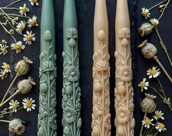 May include: Four decorative taper candles in shades of green and beige, intricately carved with floral designs. The candles are arranged on a dark slate surface, with dried flowers and seed pods scattered around them. The candles are approximately 10 inches tall.