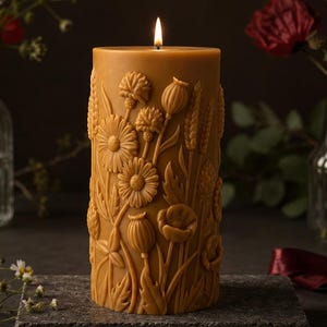 Puede incluir: Una vela de pilar encendida, de color marrón claro, con un diseño floral, que presenta margaritas, amapolas y espigas de trigo. La vela está sobre una piedra gris, con jarrones de flores en el fondo. La llama de la vela es brillante.