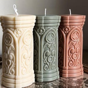 May include: Three pillar candles in cream, sage green, and dusty rose colours. Each candle features a cylindrical shape with intricate embossed architectural details, including arched patterns, floral motifs, and vertical columns, topped with a white wick.