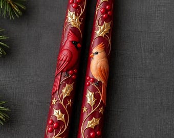 May include: Two deep red taper candles featuring raised relief designs of gold holly leaves, red berries, and small stars. One candle displays a bright red cardinal bird, while the other features a light orange cardinal bird perched on the vines.