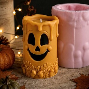 Puede incluir: Una vela cil&iacute;ndrica amarilla que presenta una cara de fantasma tallada con una expresi&oacute;n sonriente y una base decorada con hojas de roble y bellotas, colocada junto a un molde de silicona rosa a juego para fundir la forma de la vela.