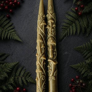May include: Two tapered candles in a muted olive green color, featuring intricate, raised relief designs of various mushrooms, winding vines, and curled fern fronds covering the entire surface of each candle, with a single white wick at the top.