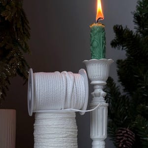 Könnte beinhalten: Eine weiße Garnrolle, die auf einer weiteren weißen Garnrolle gestapelt ist, neben einem weißen Kerzenständer mit einer brennenden grünen Kerze, die ein florales Reliefmuster aufweist. Die Kerze hat eine helle orangefarbene und gelbe Flamme.