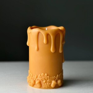Puede incluir: Una vela cil&iacute;ndrica de color amarillo dorado c&aacute;lido, con una banda decorativa en relieve de hojas de roble y bellotas en la base. El borde superior de la vela presenta un efecto de cera derretida que gotea con una mecha blanca centrada.