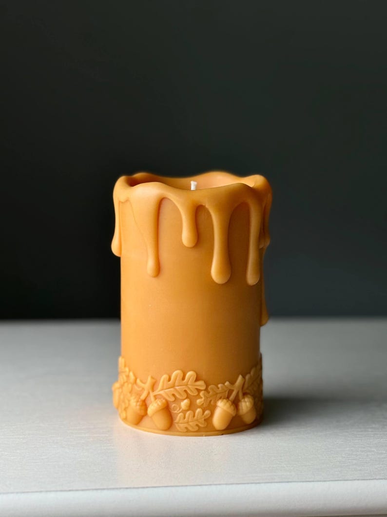 Puede incluir: Una vela cil&iacute;ndrica de color amarillo dorado c&aacute;lido, con una banda decorativa en relieve de hojas de roble y bellotas en la base. El borde superior de la vela presenta un efecto de cera derretida que gotea con una mecha blanca centrada.