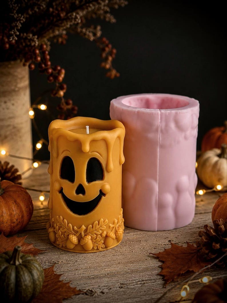 Puede incluir: Una vela cil&iacute;ndrica amarilla que presenta una cara de fantasma tallada con una expresi&oacute;n sonriente y una base decorada con hojas de roble y bellotas, colocada junto a un molde de silicona rosa a juego para fundir la forma de la vela.