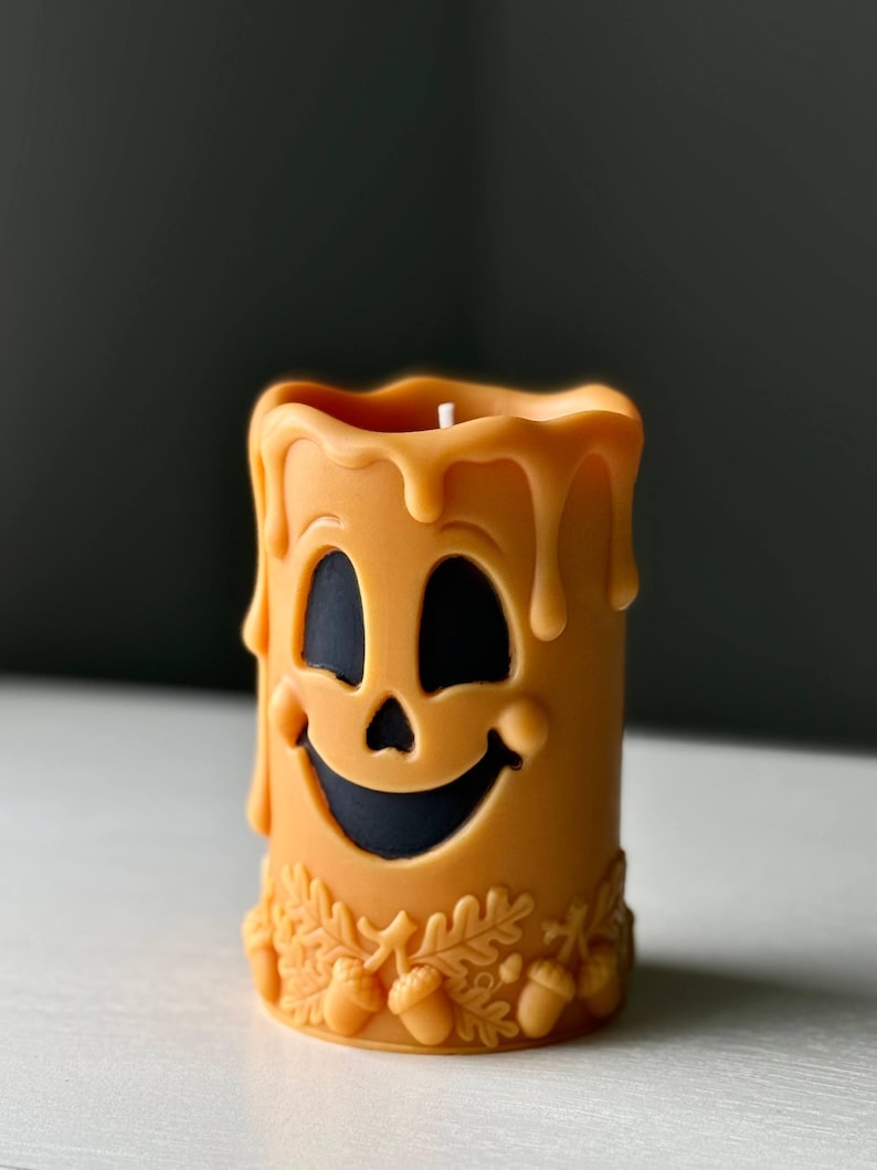 Puede incluir: Una vela cil&iacute;ndrica de cera naranja con una cara sonriente tallada y recortes negros en ojos y boca. El borde superior tiene un efecto de cera derretida y la base est&aacute; decorada con un relieve de peque&ntilde;as hojas de roble y bellotas en cera.