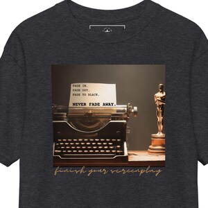 May include: Dark grey t-shirt with a vintage typewriter, a golden Oscar statue, and the text "finish your screenplay." The image on the shirt includes the text "Fade In, Fade Out, Fade to Black, Never Fade Away."