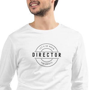 May include: A white long-sleeved shirt featuring a circular black graphic. The graphic includes the word "DIRECTOR" in bold, with "LET'S GO AGAIN" and "FOR SAFETY" arched above and below. The shirt is made of a soft material.