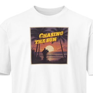 May include: White t-shirt with a square graphic depicting a sunset scene. The graphic includes palm trees, a person, and the text "Chasing The Sun" in yellow. The shirt has short sleeves and a crew neck.