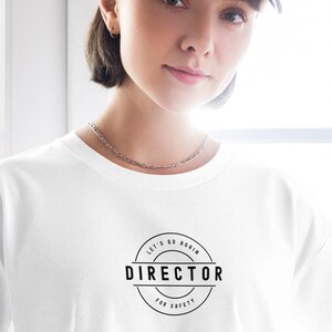 May include: White t-shirt featuring a black circular graphic with the text "Director for Safety". The shirt is paired with a denim baseball cap and a silver necklace. The text is in a bold, sans-serif font.