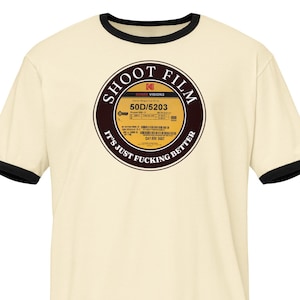 May include: A cream-coloured t-shirt with black trim around the collar and sleeves. The shirt displays a circular graphic, reminiscent of a film reel, with the text "SHOOT FILM" and "IT'S JUST FUCKING BETTER". The graphic includes film details.