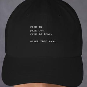May include: Black baseball cap with a curved brim. The cap features white text that reads "FADE IN. FADE OUT. FADE TO BLACK. NEVER FADE AWAY." The cap is worn by a person.