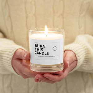 May include: A lit candle in a clear glass jar. The candle has a white label with the text "BURN THIS CANDLE" and the phrase "AFTER YOU TYPE FADE TO BLACK, BUT BEFORE YOU CLICK 'SEND'". The candle is held in front of a cream-colored sweater.