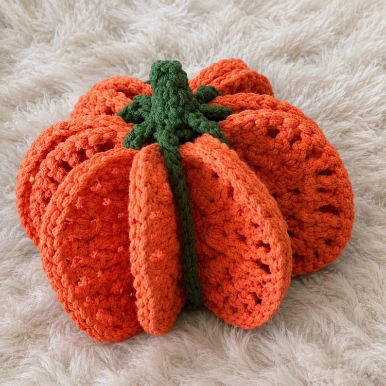 May include: A crocheted pumpkin decoration featuring vibrant orange yarn segments with a textured, open-stitch pattern. The top of the pumpkin is accented with a dark green crocheted stem and leaf detail, creating a rounded, three-dimensional shape.