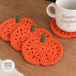 May include: Three orange pumpkin-shaped crochet coasters with green stems, arranged in a row. A white ceramic mug with the text Karen Crochet and a heart icon sits on a fourth matching coaster. A circular white label displays the text CROCHET PATTERN PDF.